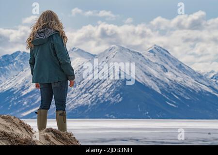 Blick von hinten auf eine Frau, die auf den Sanddünen steht und auf den gefrorenen Bennett Lake und die schneebedeckten Berge um Car blickt... Stockfoto