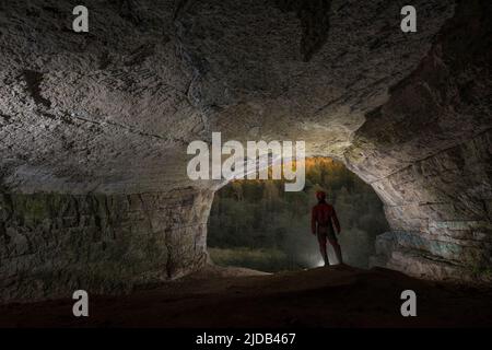 Kurz vor Sonnenuntergang steht ein Mann mit Blick auf einen Wald am Eingang zur Ignatievskaja-Höhle im Ural; Russland Stockfoto