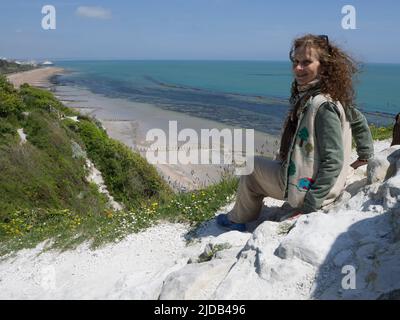 Frau sitzt auf weißen Felsen auf einem Kamm über dem Strand und der Küste und blickt auf die Kamera zurück; Eastbourne, East Sussex, England Stockfoto