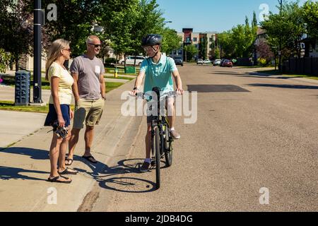 Ein junger Mann, der bereit ist, mit dem Fahrrad eine Wohnstraße hinunter zu fahren und mit seinen Eltern zu sprechen, die auf dem Bürgersteig stehen Stockfoto