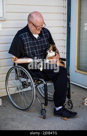 Ein Mann mit zwei Gliedmaßen, der vor seinem Haus sitzt, mit seiner Katze auf dem Schoß Albert, Alberta, Kanada Stockfoto