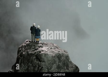 Miniatur Menschen Spielzeug konzeptuelle Fotografie. Älteres Paar, das bei dunklem bewölktem Regen über dem Bergfelsen steht, über die Zukunft nachdenkt, lebt, Stockfoto