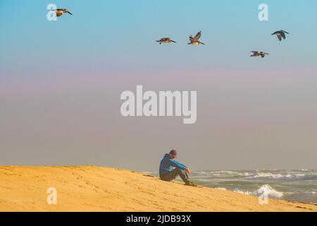 Nachdenklicher reifer Mann, der bei Sonnenuntergang am Strand am Meer sitzt Stockfoto