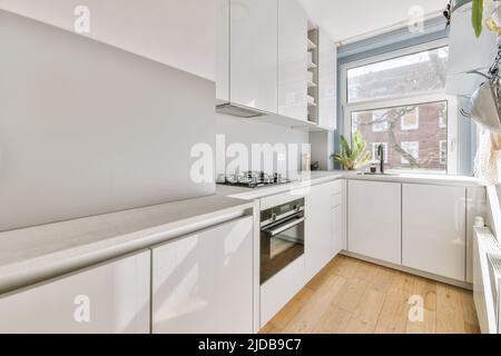 Helle kleine Wohnküche mit großem Fenster, ausgestattet mit Schränken und Regalen mit Geräten in moderner urbaner Wohnung Stockfoto