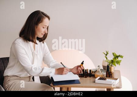 Das Aromatherapeutin-Mädchen sitzt in ihrem Büro und hält eine Flasche aromatisches Öl in den Händen und schreibt etwas auf. Es gibt ätherisches Öl Stockfoto