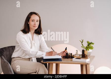 Das Aromatherapeutin-Mädchen sitzt in ihrem Büro und hält eine Flasche aromatisches Öl in den Händen und schreibt etwas auf. Es gibt ätherisches Öl Stockfoto