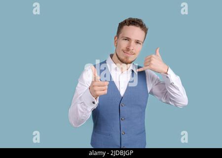 Bärtiger Geschäftsmann, der eine Anrufgeste in einem weißen Hemd und einer blauen Weste auf blauem Hintergrund im Studio zeigt, Geschäftskonzept Stockfoto