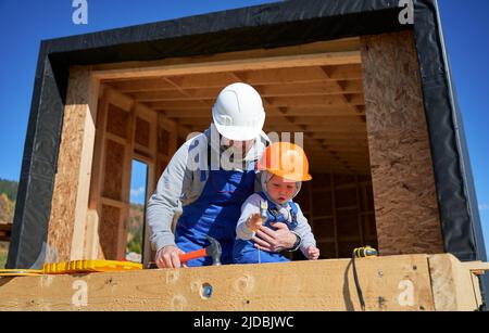 Vater mit Kleinkind Sohn Gebäude Holzrahmen Haus. Männliche Bauherren, die auf der Baustelle Nagel in die Planke schlagen, tragen an sonnigen Tagen Helm und blaue Overalls. Zimmerei- und Familienkonzept. Stockfoto