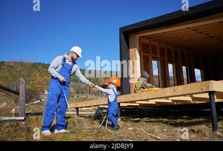 Vater mit Kleinkind Sohn Gebäude Holzrahmen Haus auf Pfahlgründung. Junge hilft seinem Vater, spielt mit Maßband auf der Baustelle, trägt Helm und blaue Overalls. Schreinerei-Konzept. Stockfoto