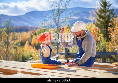 Vater mit Kleinkind Sohn Gebäude Holzrahmen Haus. Der männliche Baumeister gab dem Kind auf der Baustelle hohe fünf und trug am sonnigen Tag Helm und blaue Overalls. Zimmerei- und Familienkonzept. Stockfoto