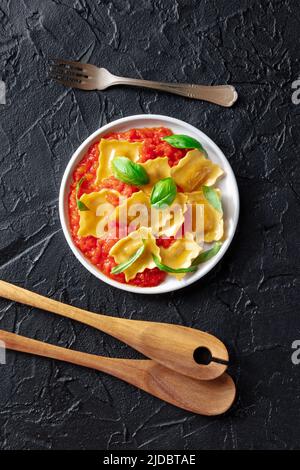 Ravioli mit Tomatensauce und frischen Basilikumblättern auf einem Teller, darüber flach auf einem schwarzen Schieferboden mit Holzlöffeln Stockfoto