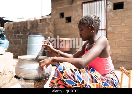 Kleines schwarzes Mädchen, das vor dem Herd in einer typisch afrikanischen Außenküche sitzt und damit beschäftigt ist, Reis zu kochen Stockfoto