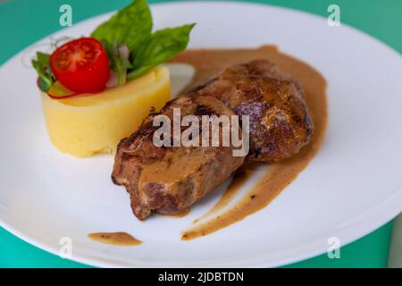 Fleischbällchen mit Kartoffelpüree auf einem weißen Teller. Fleisch in saurer Rahmsoße. Tomaten und Basilikum. Essen im Restaurant. Mittagspause. Medaillons. Stockfoto