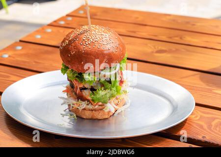 Großer großer, leckerer Hamburger oder Rinderburger mit Gemüse und Sauce auf Metallplatte auf Holztisch. Brunch an sonnigen Tagen. Nahaufnahme, Platz kopieren, fro Stockfoto