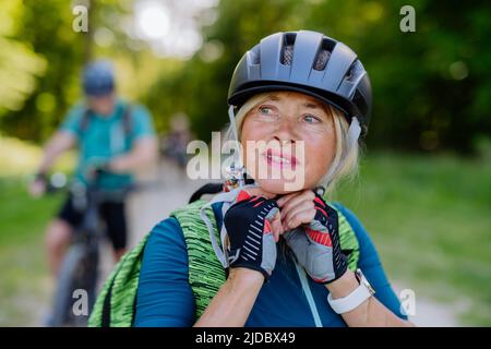 Aktives Seniorenpaar auf Fahrrädern im Sommerpark, Frau setzt Helm, gesundes Lifestyle-Konzept. Stockfoto