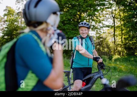 Aktives Seniorenpaar auf Fahrrädern im Sommerpark, Frau setzt Helm, gesundes Lifestyle-Konzept. Stockfoto