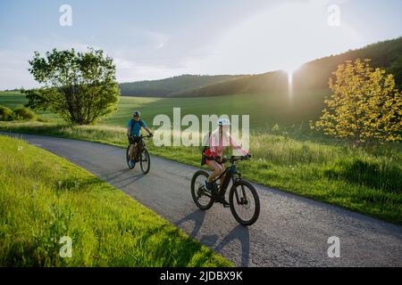 Aktives Seniorenpaar, das auf der Straße im Sommerpark Elektrofahrräder fährt, gesundes Lifestyle-Konzept. Stockfoto