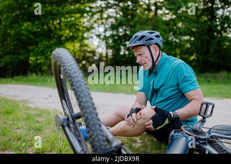 Aktiver Senior in Sportkleidung fiel vom Fahrrad auf den Boden und verletzte sich im Sommer im Park sein Knie. Stockfoto