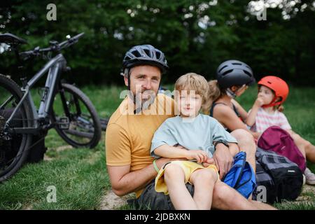 Junge Familie mit kleinen Kindern, die sich nach einer Radtour ausruhen und im Sommer auf dem Rasen im Park sitzen. Stockfoto