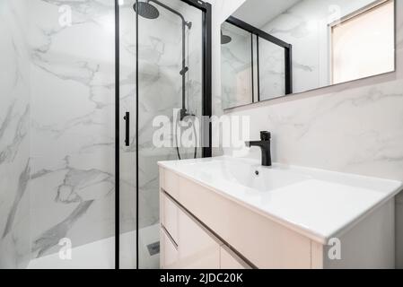 Toilette mit weißem Resin-Schrank, rahmenloser rechteckiger Spiegel, Duschkabine mit Glaswand und schwarzer Metallverkleidung und Marmorfliesen. Stockfoto