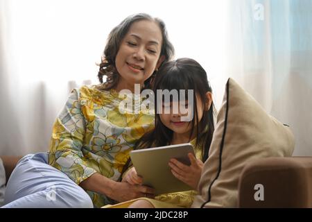 Lächelnde reife Oma und kleine Vorschulkinder nutzen digitale Tablet-Surfen im Internet und beobachten Online-Cartoon zusammen auf der Couch zu Hause Stockfoto