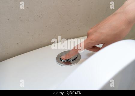 Die Hand einer Frau drückt einen Knopf auf einer weißen Toilette, um zu spülen Stockfoto