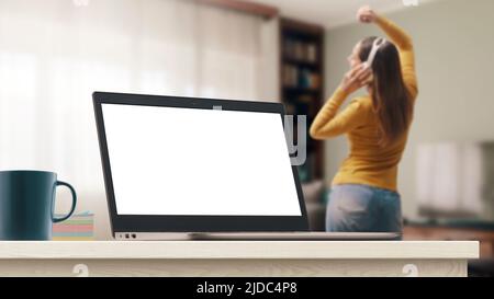 Laptop mit leerem Bildschirm und junge Frau, die zu Hause tanzt, trägt kabellose Kopfhörer und hört online Musik Stockfoto