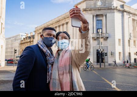 Ein Paar in der Stadt trägt Gesichtsmasken, hält das Handy und nimmt Selfie auf Stockfoto