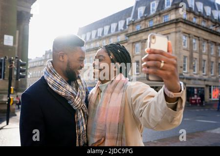 Fröhliches Paar, das in der Stadt von Angesicht zu Angesicht lächelt, Frau hält das Telefon und nimmt Selfie auf Stockfoto