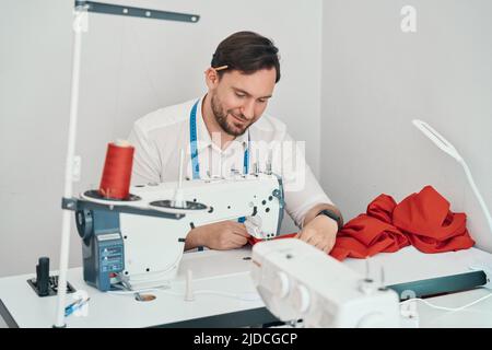 Fröhlicher männlicher Schneidermeister mit Nähmaschine in der Werkstatt Stockfoto