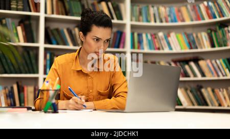 Fokussierte Frau mit Laptop und Schreiben im Notebook, Online-Studium in der Bibliothek Innenraum, Panorama mit freiem Platz Stockfoto