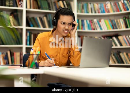 Die junge Frau, die mit einem Laptop online studiert und Hörprobleme in ihren Kopfhörern hat, sitzt im Inneren der Bibliothek Stockfoto