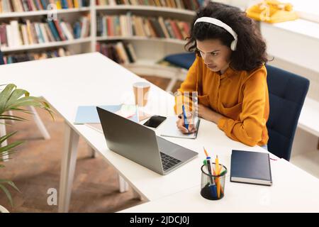 Fokussierte schwarze Frau in Kopfhörern, die Online-Unterricht auf einem Laptop macht, im Notebook schreibt, während sie in der Bibliothek sitzt Stockfoto