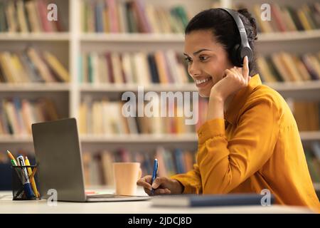 Positive gemischte Renndame, die in der Bibliothek sitzt, einen Laptop benutzt und sich Notizen macht, einen Vortrag oder ein Webinar ansieht Stockfoto