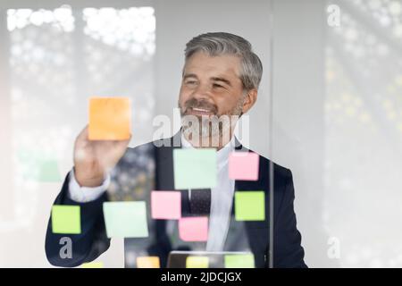 Ein glücklicher Geschäftsmann mittleren Alters, der auf klebrigen Papiernotizen an der Glaswand schrieb Stockfoto