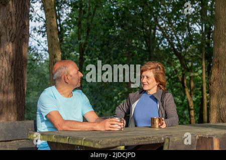 Ältere Paare, die einander zuschauen und lächeln, während sie auf einem Picknick-Holztisch sitzen, der im Wald ruht Stockfoto
