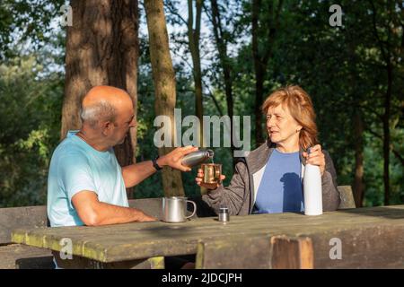 Senior Mann gießt Wasser aus einer wiederverwendbaren Flasche in einen metallischen Becher, während er im Park sitzt Stockfoto