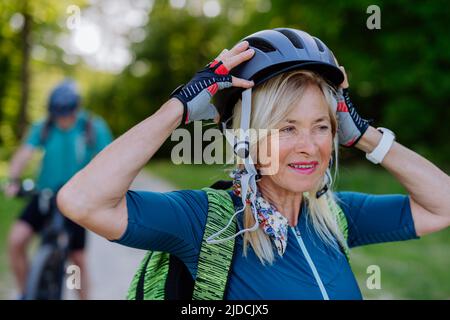 Aktives Seniorenpaar auf Fahrrädern im Sommerpark, Frau setzt Helm, gesundes Lifestyle-Konzept. Stockfoto
