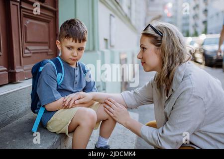 Mutter tröstete ihren kleinen Sohn an seinem ersten Schultag, saß auf der Treppe und sagte vor der Schule Auf Wiedersehen. Stockfoto