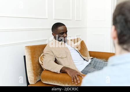 Ein schwarzer männlicher Patient, der sich einer Psychotherapiesitzung mit einem Berater in einer psychiatrischen Klinik unterziehen muss. Junger Mann mit emotionalen Problemen konsultiert Stockfoto