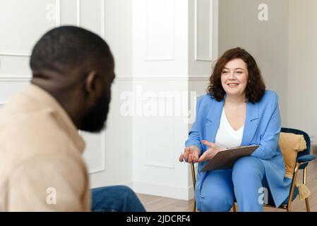 Eine Psychologin führt mit einem afroamerikanischen Mann eine psychologische Therapie durch, notiert sich in einem Clipboard. Stockfoto