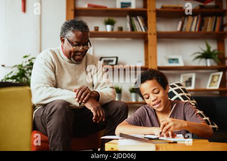 Ein afroamerikanischer Großvater sitzt zu Hause mit seinem Enkel und hilft ihm bei den Hausaufgaben. Stockfoto