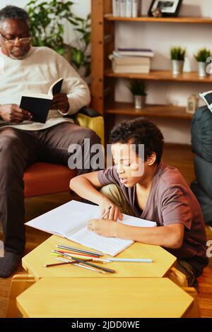 Ein afroamerikanischer Großvater sitzt auf seinem Stuhl und liest ein Buch, während sein geliebter Enkel Hausaufgaben macht. Idyllischer Nachmittag bei den Großeltern. Stockfoto