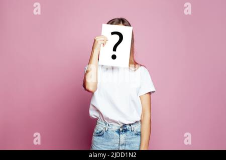 Hübsche Frau, die ihr Gesicht mit einem weißen Papierbogen mit einem gezeichneten Fragezeichen bedeckt, wie eine Maske, um ihre Identität auf einem rosa Hintergrund zu verbergen Stockfoto