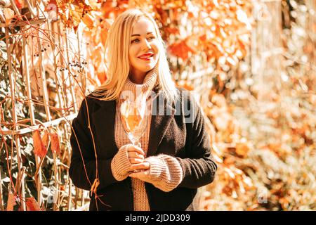 Die schöne blonde 35-Jährige in blackem Kantel und beigefarbenem Pullover hält Glas mit Herbstcocktail in den Händen und genießt einen sonnigen Tag im Stadtpark. Farben Stockfoto