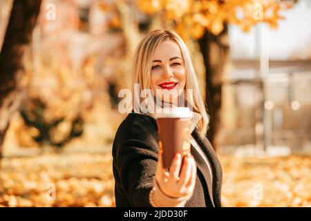 Charmantes Blondine mit leuchtend rotem Lippenstift in schwarzem Kaschmirmantel und Strickwollpullover lädt am Herbsttag zum leckeren heißen Kaffee ein Stockfoto