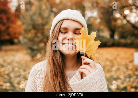 Charmante junge Frau in Strickmütze und Pullover lächelt glücklich auf Spaziergang im Herbstpark, halten gelbe Ahornblatt vor ihr. Genießen Sie den Herbst Stockfoto