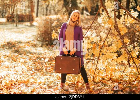 Fröhliche Frau in kuscheliger Kleidung mit altem Koffer in den Händen posiert im Herbstpark voller gefallener Blätter. Herbstfarben. Erholung im Park Stockfoto