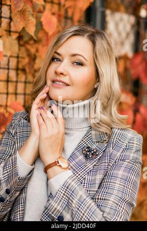 Porträt einer gepflegten, charmanten Blondine von 35-40 Jahren mit langen Haaren an der Wand aus roten Herbstblättern im Stadtgarten am sonnigen Herbsttag Stockfoto