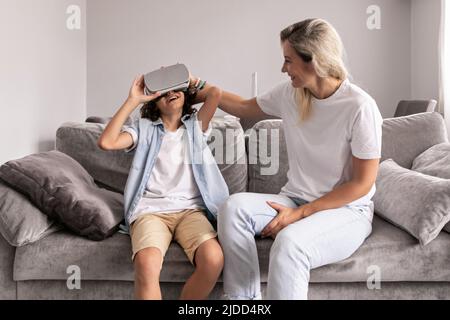 Mutter hilft seinem Sohn, im Wohnzimmer zu Hause eine Virtual-Reality-Headset-Brille zu tragen und Spaß zu haben Stockfoto